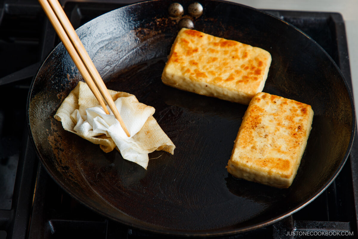 Two rectangular pieces of tofu are being pan-fried on a skillet. A hand holding chopsticks is wiping the pan with a paper towel, showing part of the cooking process.