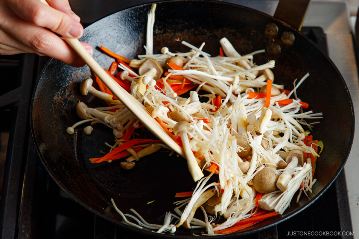 A hand uses chopsticks to stir-fry sliced carrots and a variety of mushrooms, including enoki and shimeji, in a black pan on a stovetop.