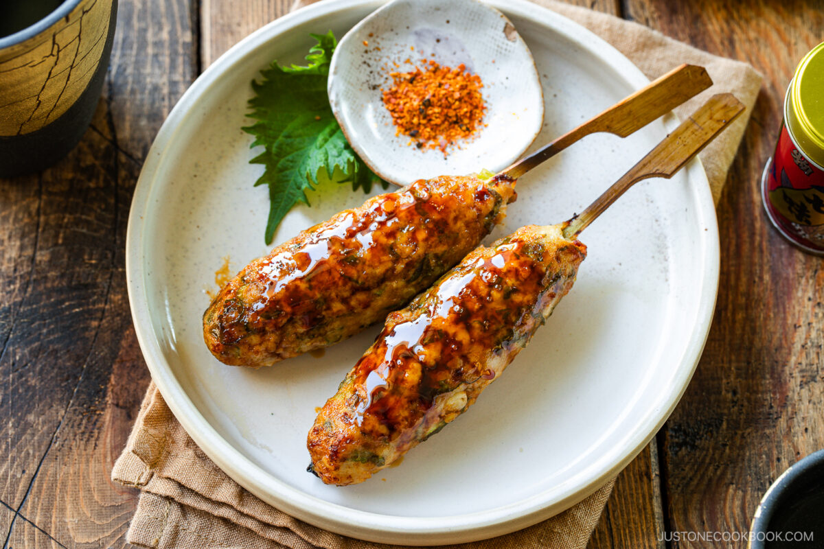 Two grilled chicken tsukune skewers glazed with sauce are served on a white plate with a green shiso leaf and a small dish of red seasoning. The plate rests on a tan cloth and wooden table.