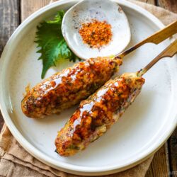 Two grilled chicken tsukune skewers glazed with sauce are served on a white plate with a green shiso leaf and a small dish of red seasoning. The plate rests on a tan cloth and wooden table.