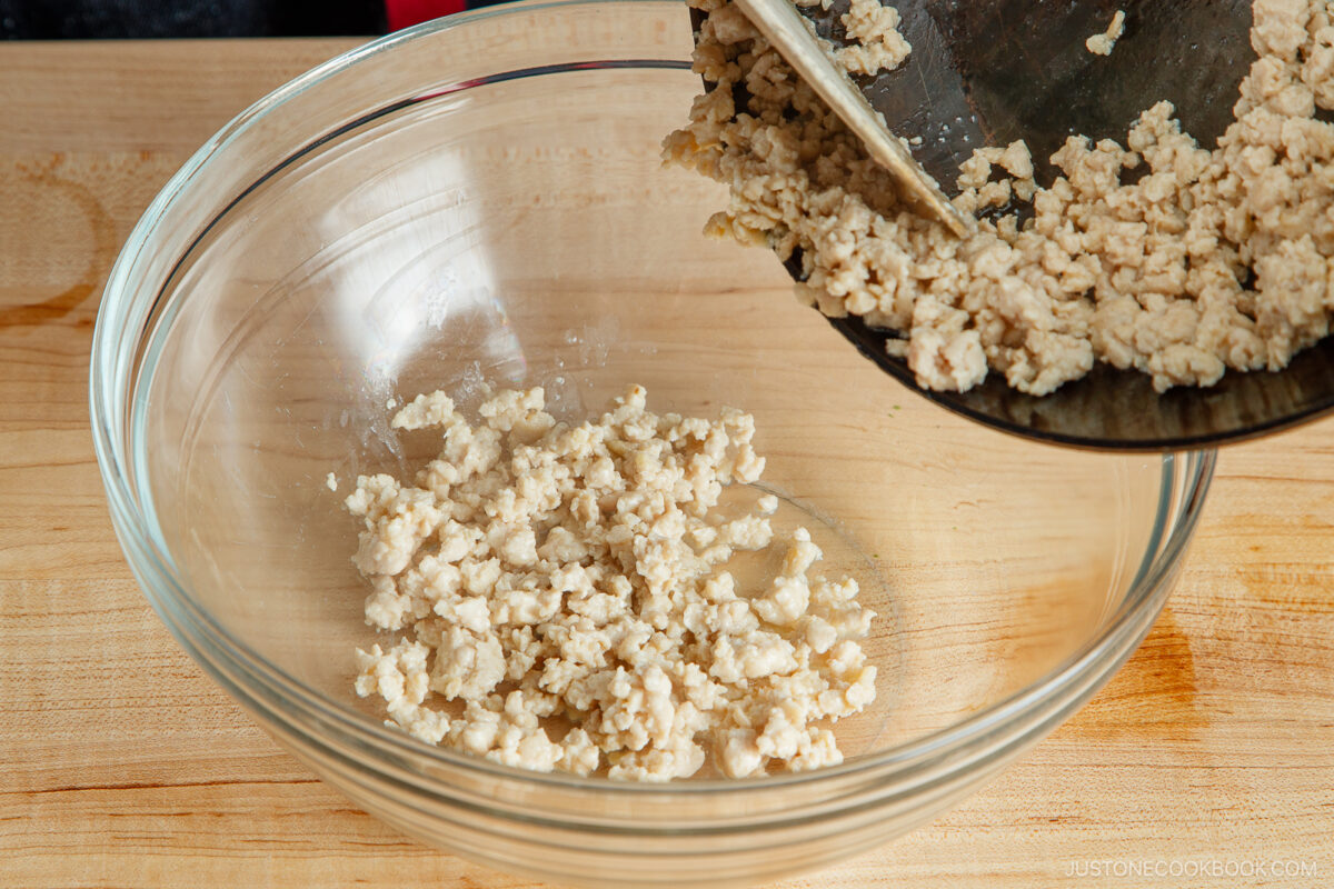 Cooked ground turkey being poured from a skillet into a clear glass mixing bowl on a wooden countertop.
