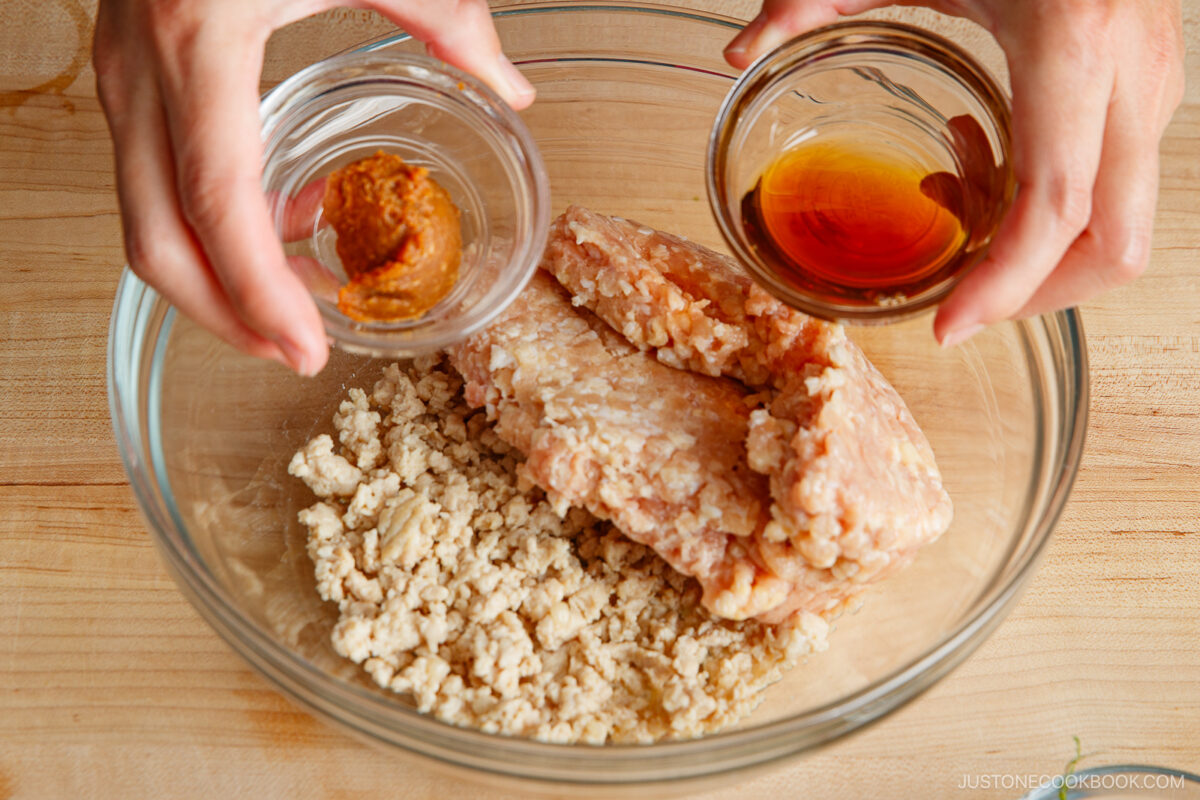 A person holds small bowls of miso paste and soy sauce above a glass bowl containing raw ground chicken on a wooden surface, preparing to add the ingredients.