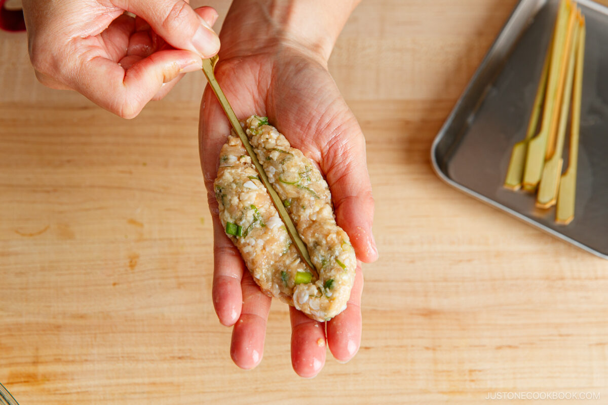 A hand shapes seasoned minced meat around a lemongrass stalk, preparing it for grilling, with additional lemongrass stalks on a tray nearby on a wooden surface.