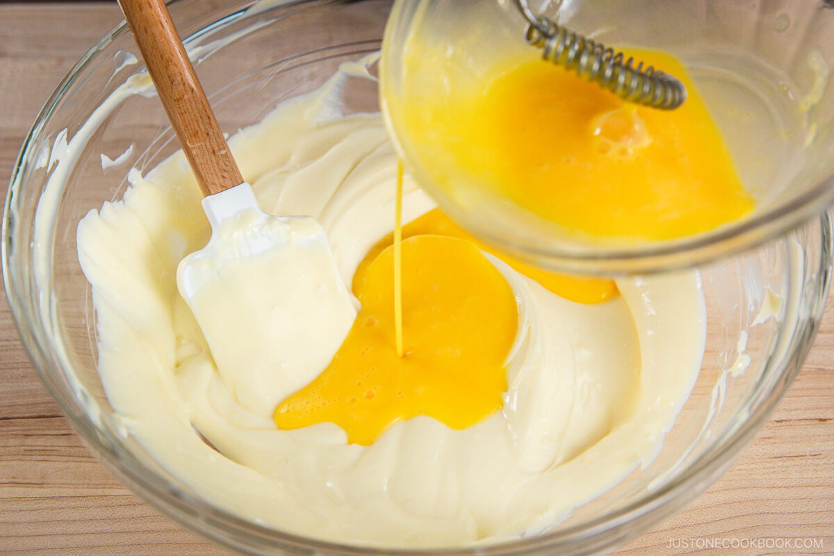 A glass bowl of creamy white mixture is being combined with whisked eggs poured from another container, with a spatula resting in the bowl on a wooden surface.