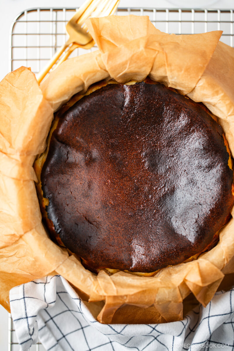 A close-up of a burnt Basque cheesecake with a dark, caramelized top, sitting in parchment paper on a wire rack. A white kitchen towel and golden spatula are partially visible beside the cheesecake.