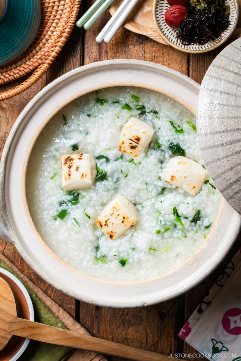 A bowl of rice porridge with grilled tofu cubes and greens, served in a ceramic pot on a wooden table with chopsticks, side dishes, and a wooden spoon nearby.