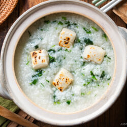 A ceramic pot filled with rice porridge topped with grilled tofu cubes and chopped greens sits on a wooden table, next to a wooden spoon and various dishes.