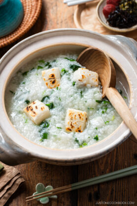 A bowl of Japanese rice porridge topped with grilled mochi cubes and chopped green onions, served in a clay pot with a wooden spoon on a wooden table. Chopsticks and small side dishes are nearby.