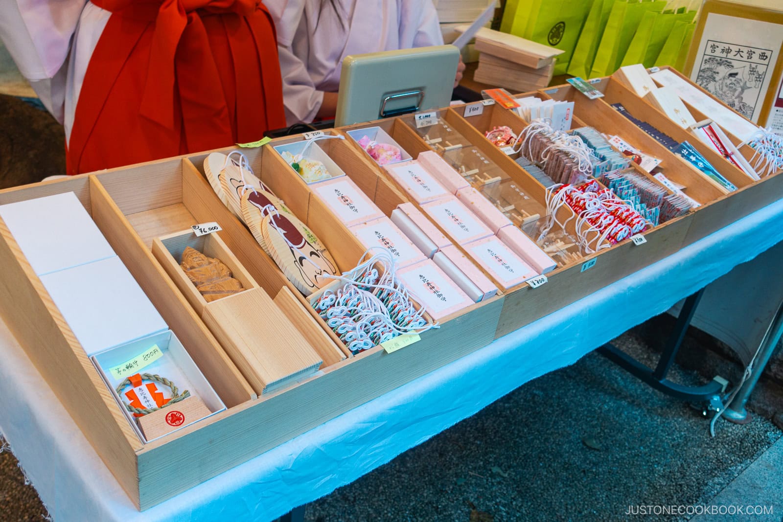 A display of various Japanese omamori (amulets) and charms neatly arranged in wooden trays on a table at a shrine. A person in traditional attire is partially visible behind the counter.