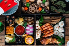 A colorful Japanese New Year’s feast (osechi-ryori) arranged in lacquered boxes, featuring shrimp, rolled omelette, black beans, pickled vegetables, fish roe, lotus root, and decorative garnishes on a wooden table.