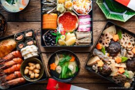 An assortment of traditional Japanese New Year foods, including colorful seafood, vegetables, and sweets arranged in lacquered boxes and bowls on a wooden table.
