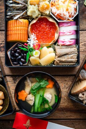 An assortment of traditional Japanese New Year foods, including colorful seafood, vegetables, and sweets arranged in lacquered boxes and bowls on a wooden table.