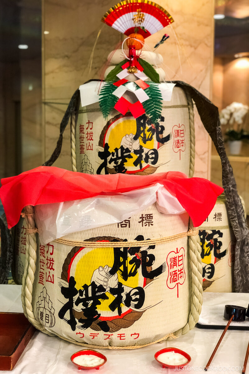 Large decorative sake barrels wrapped in straw, adorned with Japanese writing and symbols. The front barrel is open and covered with a red cloth, and festive ornaments are displayed on top. The background shows a marble wall.