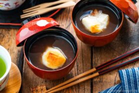 Two bowls of sweet red bean soup topped with grilled mochi sit on a wooden table, accompanied by a cup of green tea, chopsticks, and a blue-striped cloth.