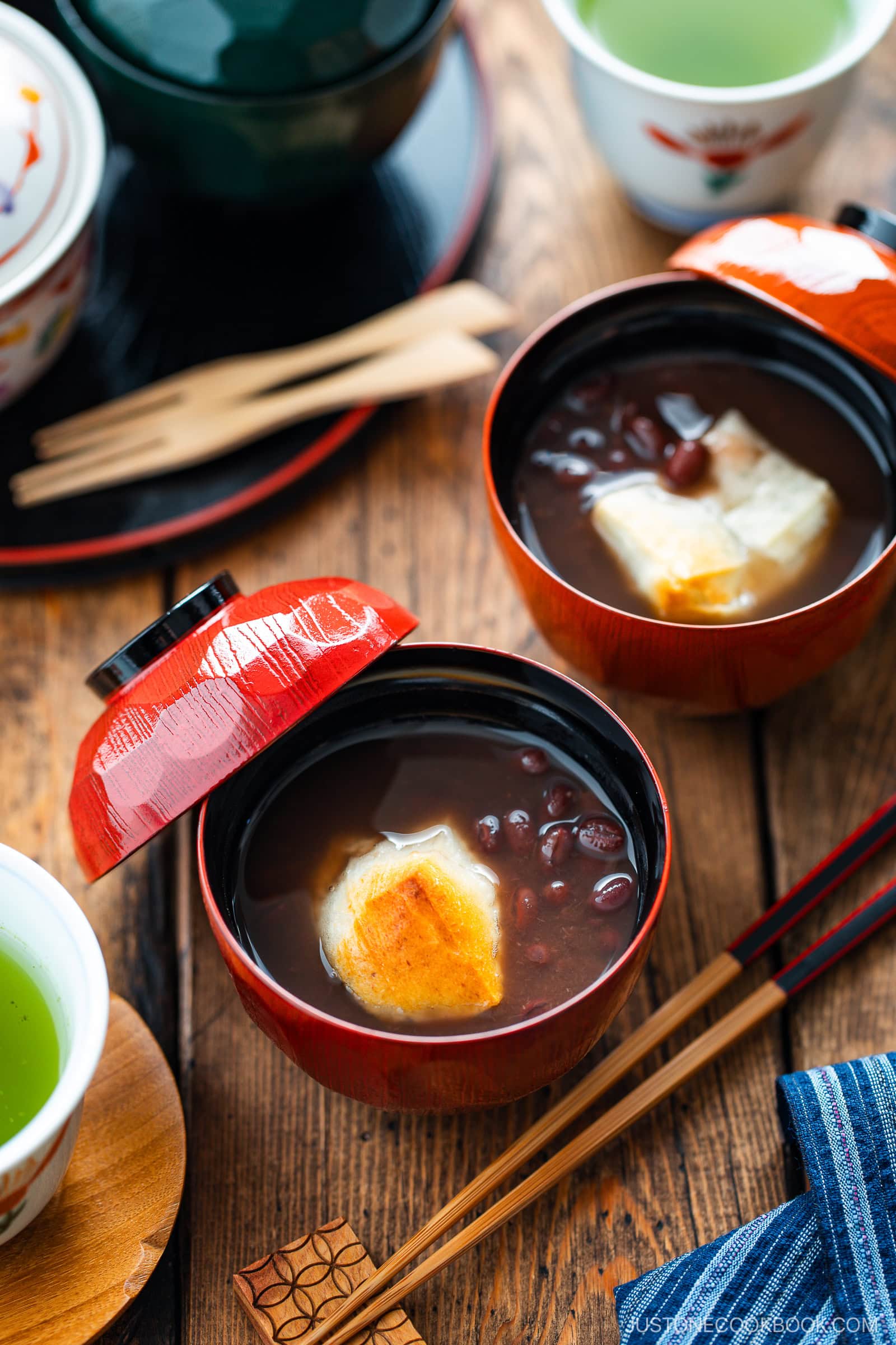 Two red lacquer bowls filled with sweet red bean soup and toasted mochi sit on a wooden table, surrounded by green tea cups, wooden chopsticks, and bamboo utensils.