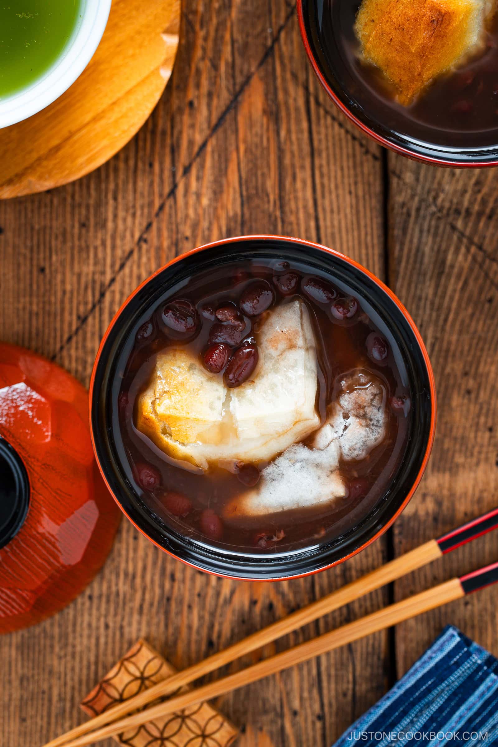 A bowl of traditional Japanese ozoni soup with grilled mochi and red beans, set on a wooden table with chopsticks nearby and part of a cup of green tea visible.