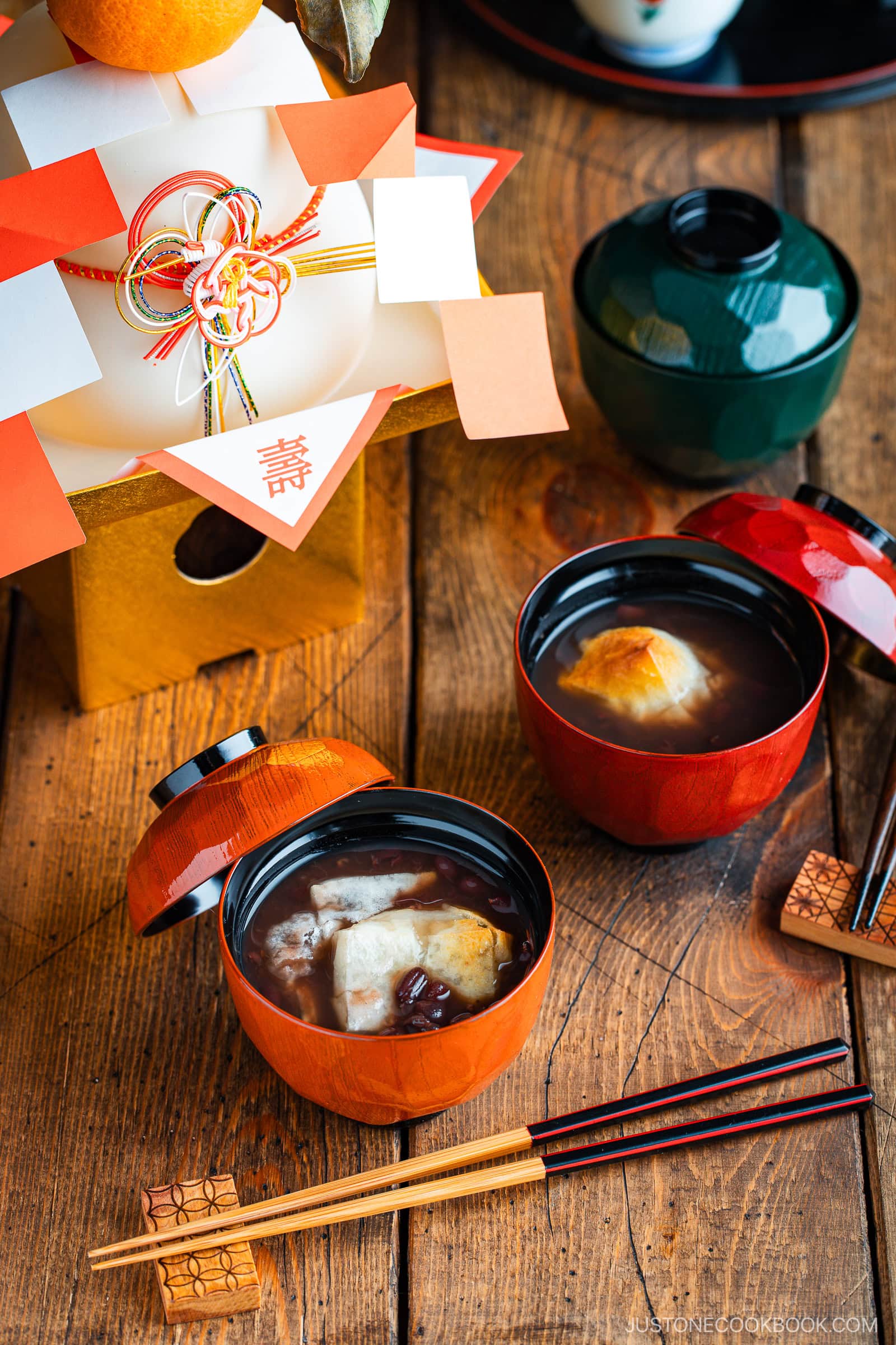 Two lacquered bowls of zenzai (sweet red bean soup with toasted mochi) sit on a wooden table with chopsticks. Nearby is a decorative kagami mochi and lidded green bowl, evoking a Japanese New Year celebration.