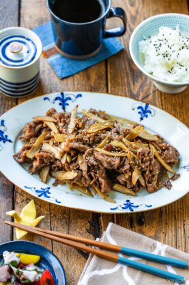 A plate of Beef and Gobo Stir Fry garnished with sesame seeds sits on a wooden table, accompanied by a bowl of white rice, a cup of tea, chopsticks, and a small salad.