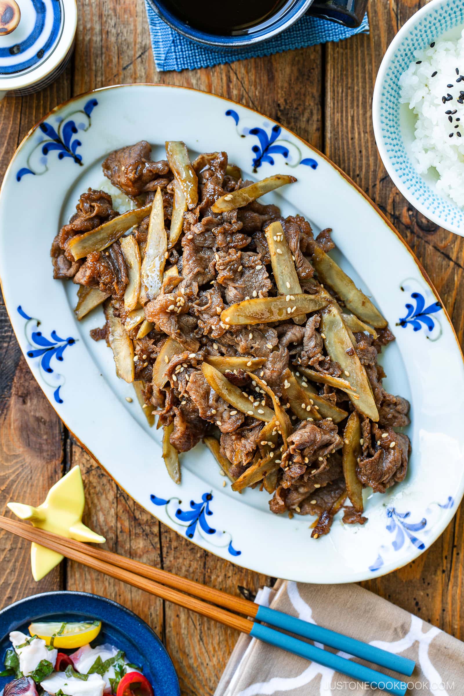 A blue-and-white oval plate holds Beef and Gobo Stir Fry—tender beef and burdock root garnished with sesame seeds. A bowl of rice, a small dish, and chopsticks with a blue holder are arranged nearby on a wooden table.