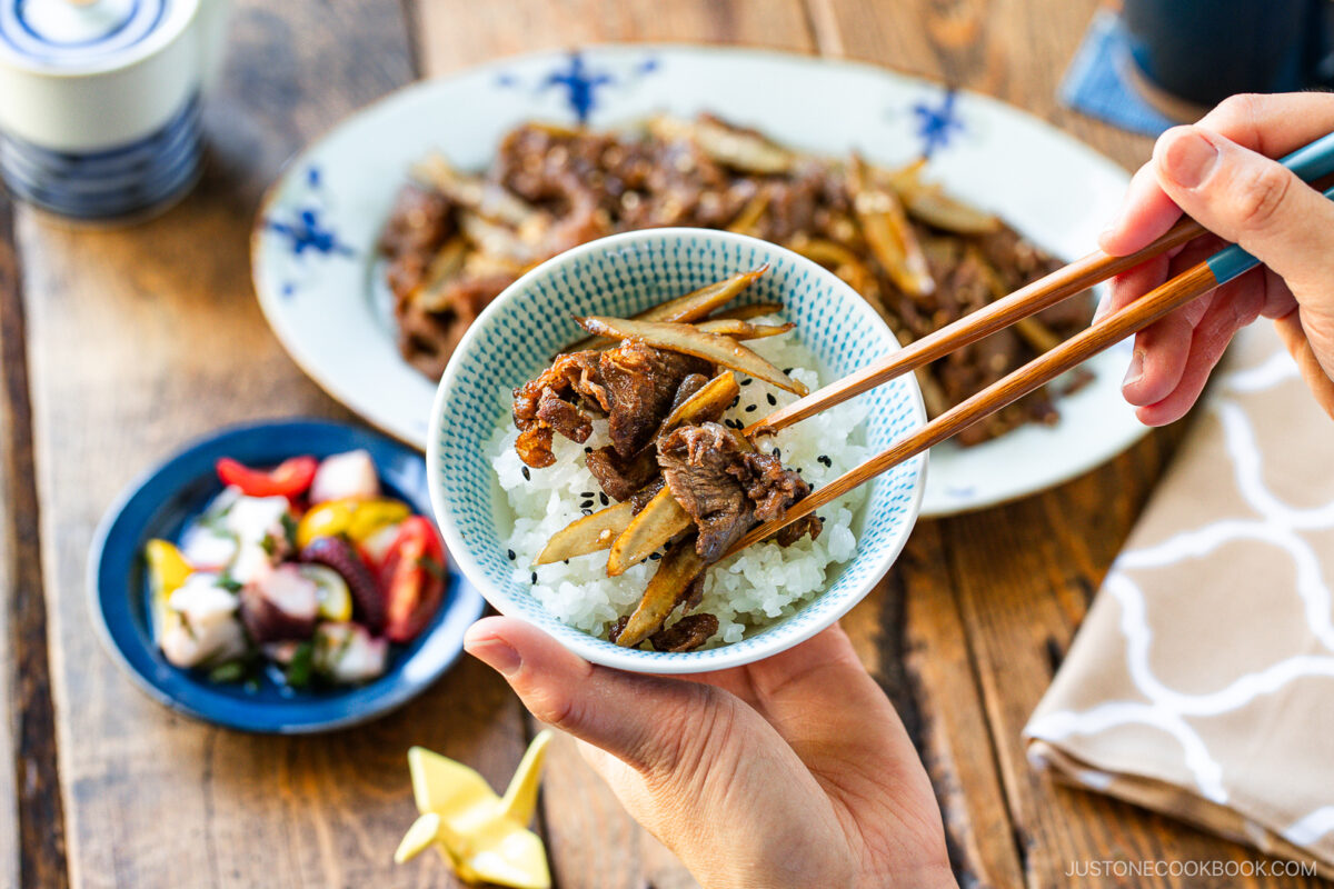 A person uses chopsticks to pick up beef and vegetables from a bowl of rice, with Beef and Gobo Stir Fry, a dish of colorful pickles, and a cup on a wooden table in the background.
