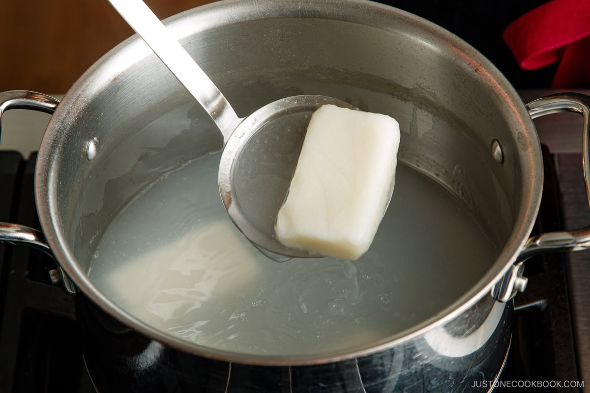 A metal pot on a stove contains water and two pieces of white mochi, with one piece being lifted out by a metal ladle.