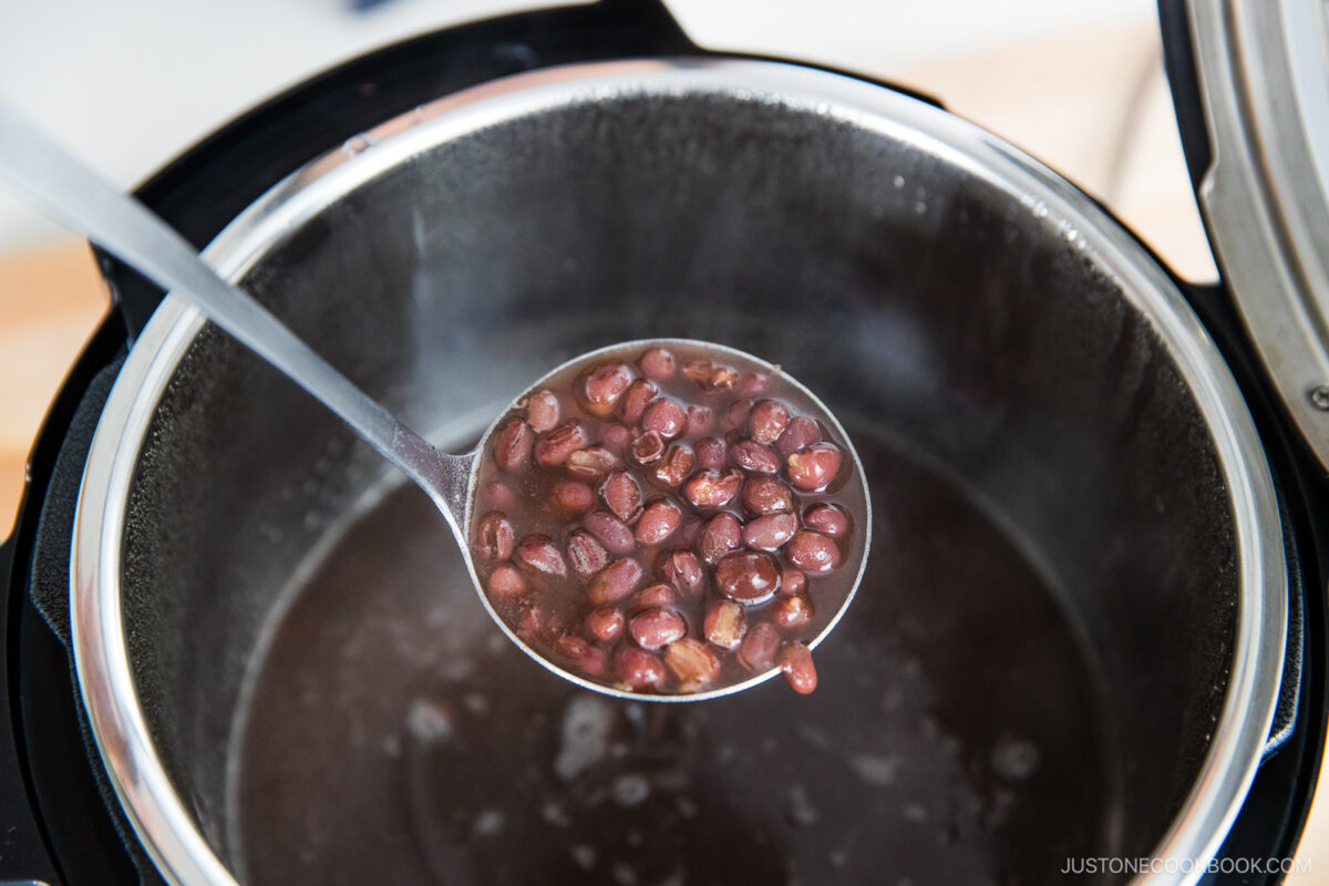 A metal ladle lifting cooked red beans from a pot filled with dark liquid, with steam rising from the hot food.