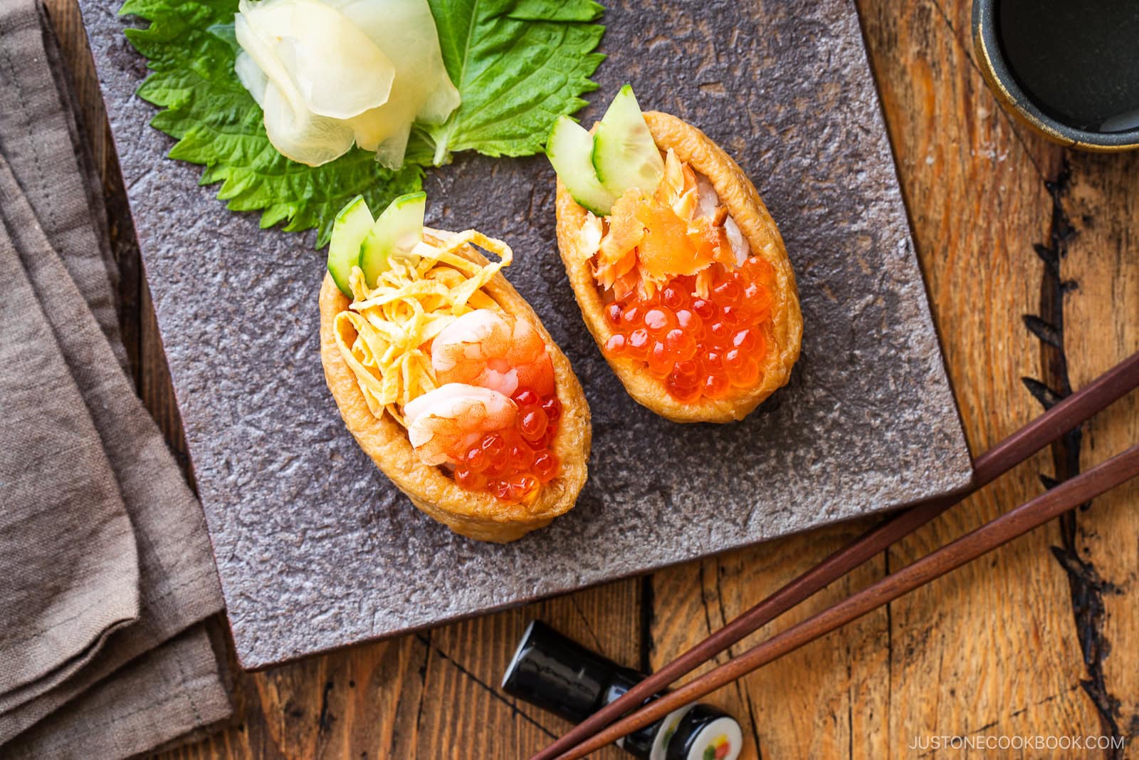 Two inari sushi pieces topped with shrimp, shredded egg, salmon roe, and cucumber slices are served on a square stone plate with pickled ginger and shiso leaf, alongside wooden chopsticks and a small soy sauce dish.