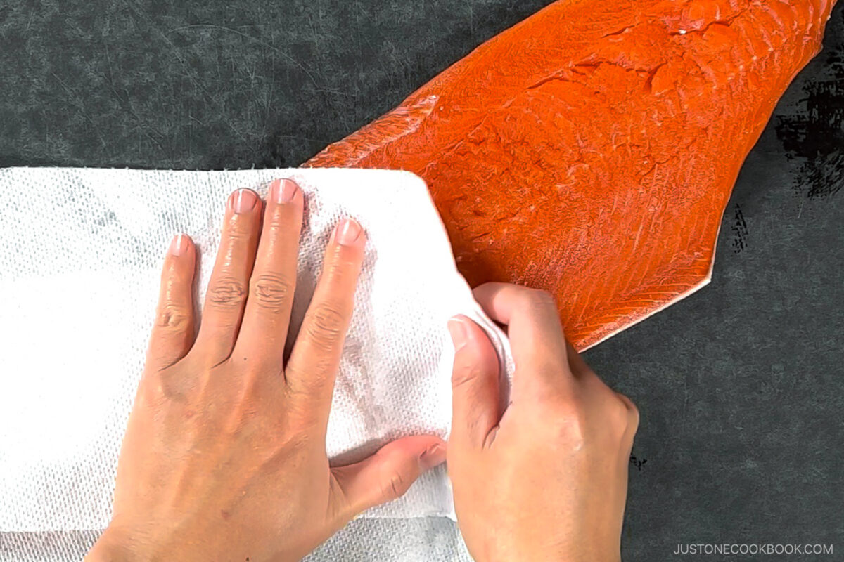 Two hands pat a raw salmon fillet dry with a paper towel on a dark surface. The salmon is partially covered by the towel as it is being prepared.
