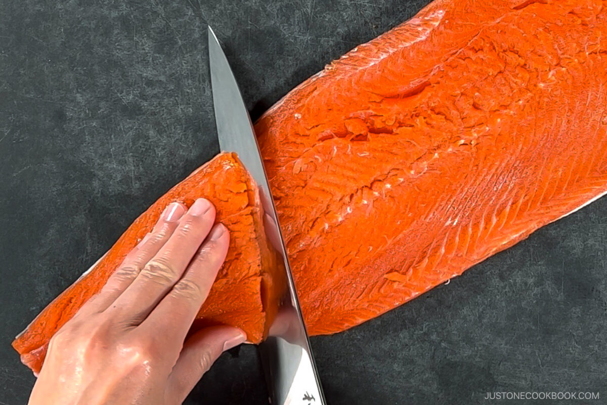 A hand uses a large knife to slice a raw salmon fillet on a dark cutting board. The fishs vibrant orange flesh and the texture of the fillet are clearly visible.