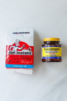 A white and red bag of Saf-Instant yeast sits next to a jar of Fleischmann’s Bread Machine Instant Yeast on a white marble surface.