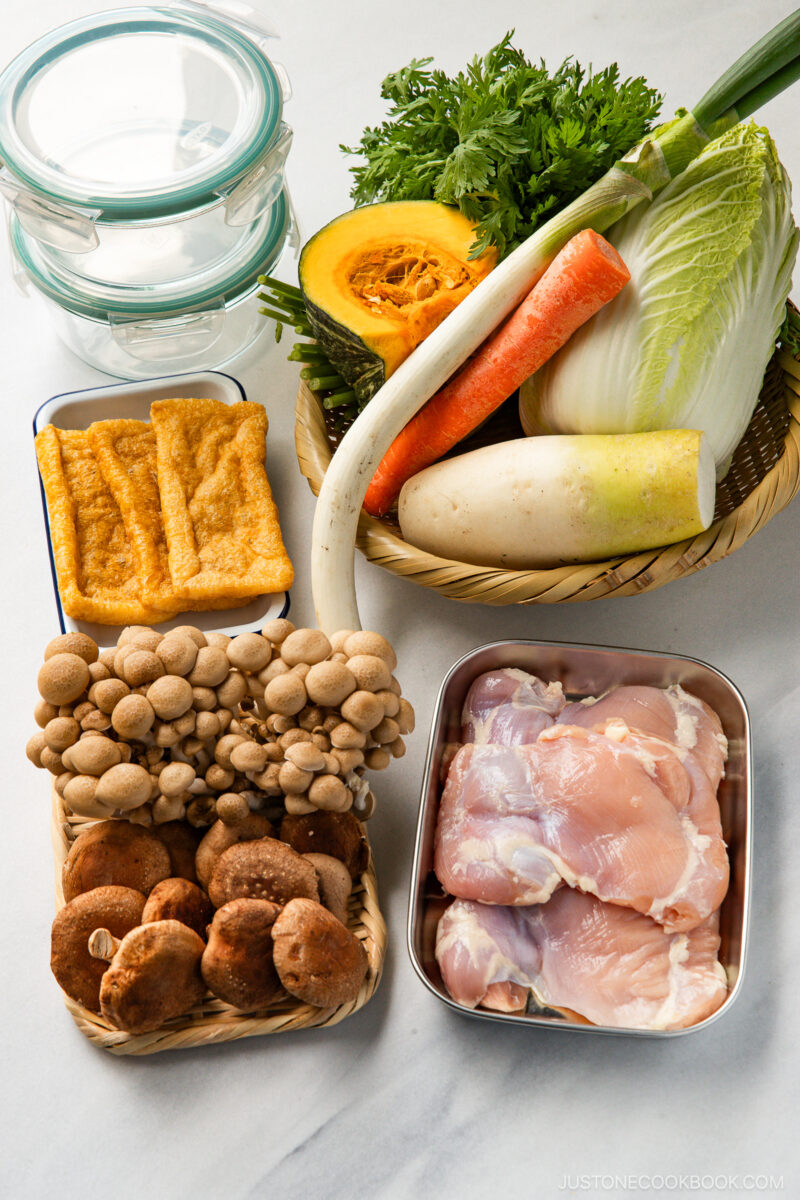 A variety of fresh ingredients on a countertop for Japanese hot pot meal prep: chicken thighs, shiitake and shimeji mushrooms, fried tofu, carrot, napa cabbage, daikon radish, kabocha squash, green onions, leafy herbs, and two glass containers.