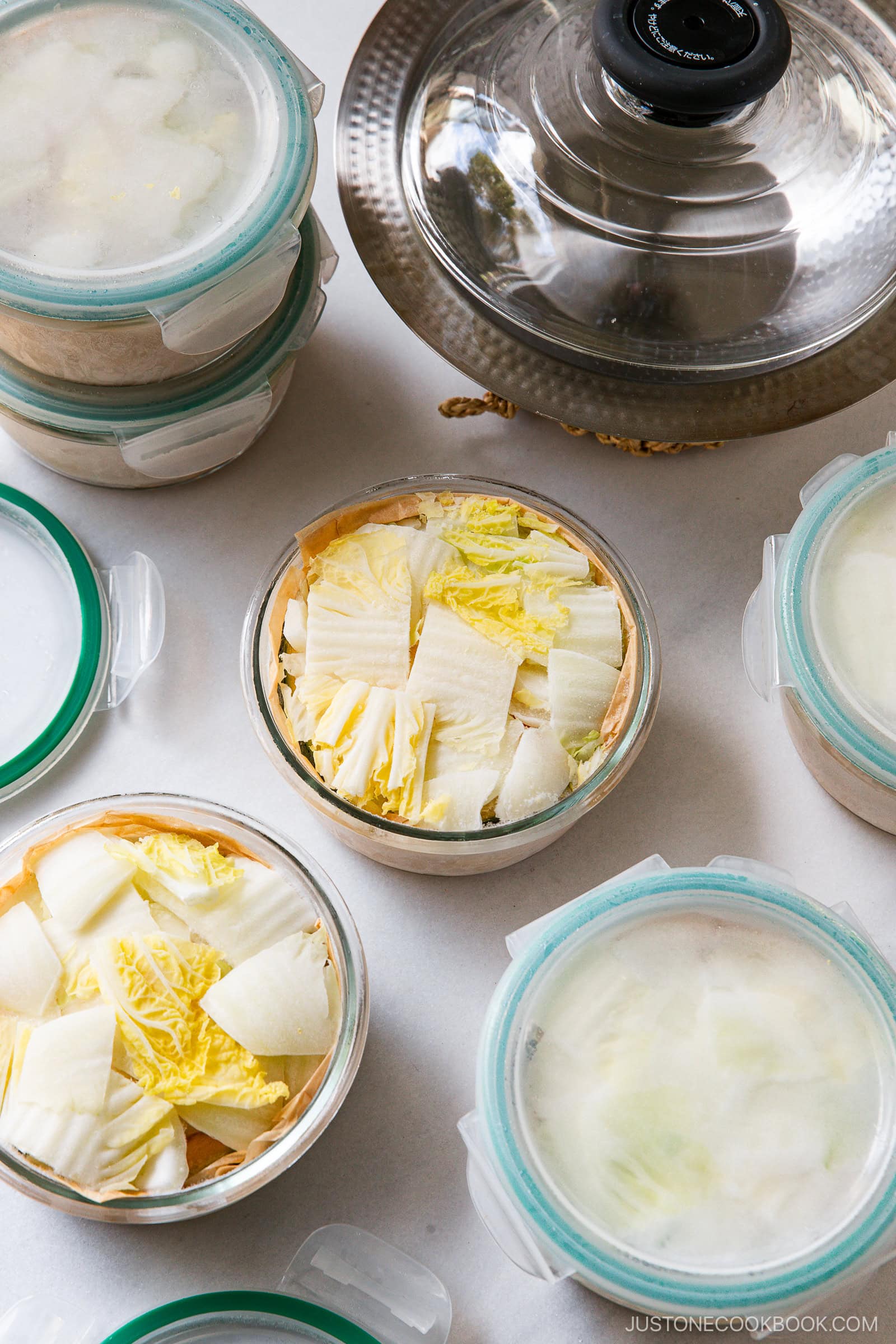 Round glass containers filled with chopped napa cabbage and other vegetables, some with lids on and one open, are arranged on a white surface next to a pot with a glass lid—perfect for Japanese hot pot meal prep.