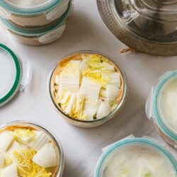 Glass containers filled with chopped napa cabbage, some with lids on and some open, are arranged on a white surface—ideal for Japanese hot pot meal prep. A metal pot with a glass lid is partially visible in the upper right corner.