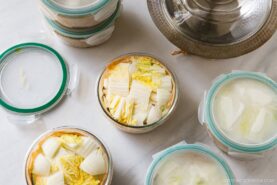 Glass containers filled with chopped napa cabbage, some with lids on and some open, are arranged on a white surface—ideal for Japanese hot pot meal prep. A metal pot with a glass lid is partially visible in the upper right corner.