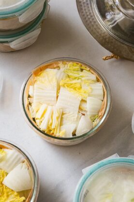 Glass containers filled with chopped napa cabbage, some with lids on and some open, are arranged on a white surface—ideal for Japanese hot pot meal prep. A metal pot with a glass lid is partially visible in the upper right corner.