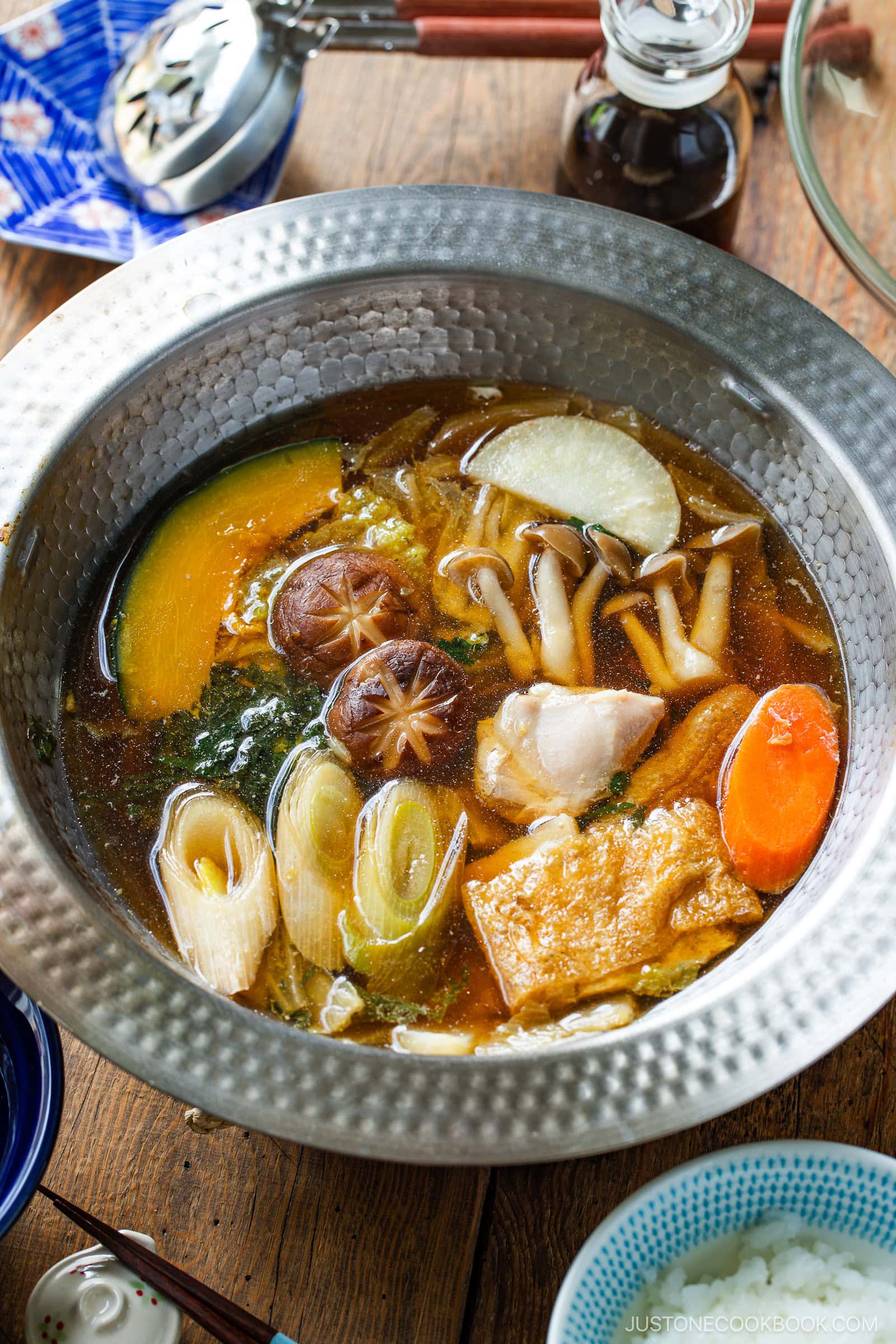 A Japanese hot pot meal prep features a steaming broth brimming with pumpkin, leek, daikon, carrot, mushrooms, tofu, and tender fish pieces. It’s served on a wooden table with chopsticks and dipping sauce nearby.
