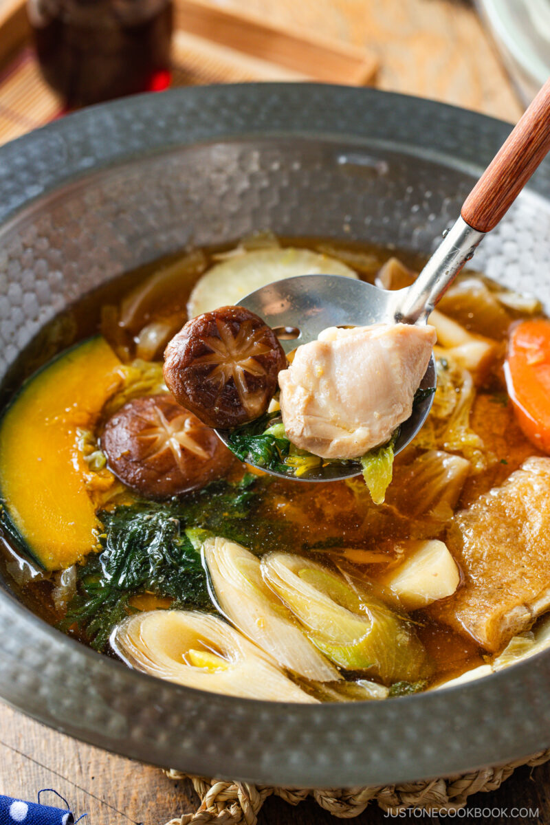 A close-up of a hearty Japanese hot pot meal prep featuring mushrooms, pumpkin, leafy greens, carrots, and leeks in a rich broth. A spoon lifts a piece of chicken and vegetables above the bowl.
