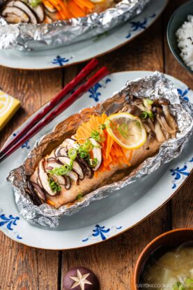 Miso salmon in foil baked to perfection, topped with sliced mushrooms, julienned carrots, and lemon, garnished with chopped green onions. Served on a decorative plate with chopsticks beside it.