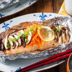 A miso salmon in foil fillet topped with sliced mushrooms, shredded carrots, lemon, and green onions, served on a decorative plate with red chopsticks and a bowl of rice nearby.