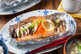 A miso salmon in foil fillet topped with sliced mushrooms, shredded carrots, lemon, and green onions, served on a decorative plate with red chopsticks and a bowl of rice nearby.