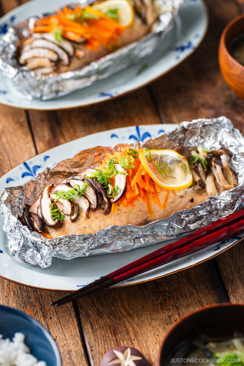Miso salmon in foil baked to perfection, topped with sliced mushrooms, shredded carrots, green onions, and lemon slices, served on a decorative plate with red chopsticks on a wooden table.
