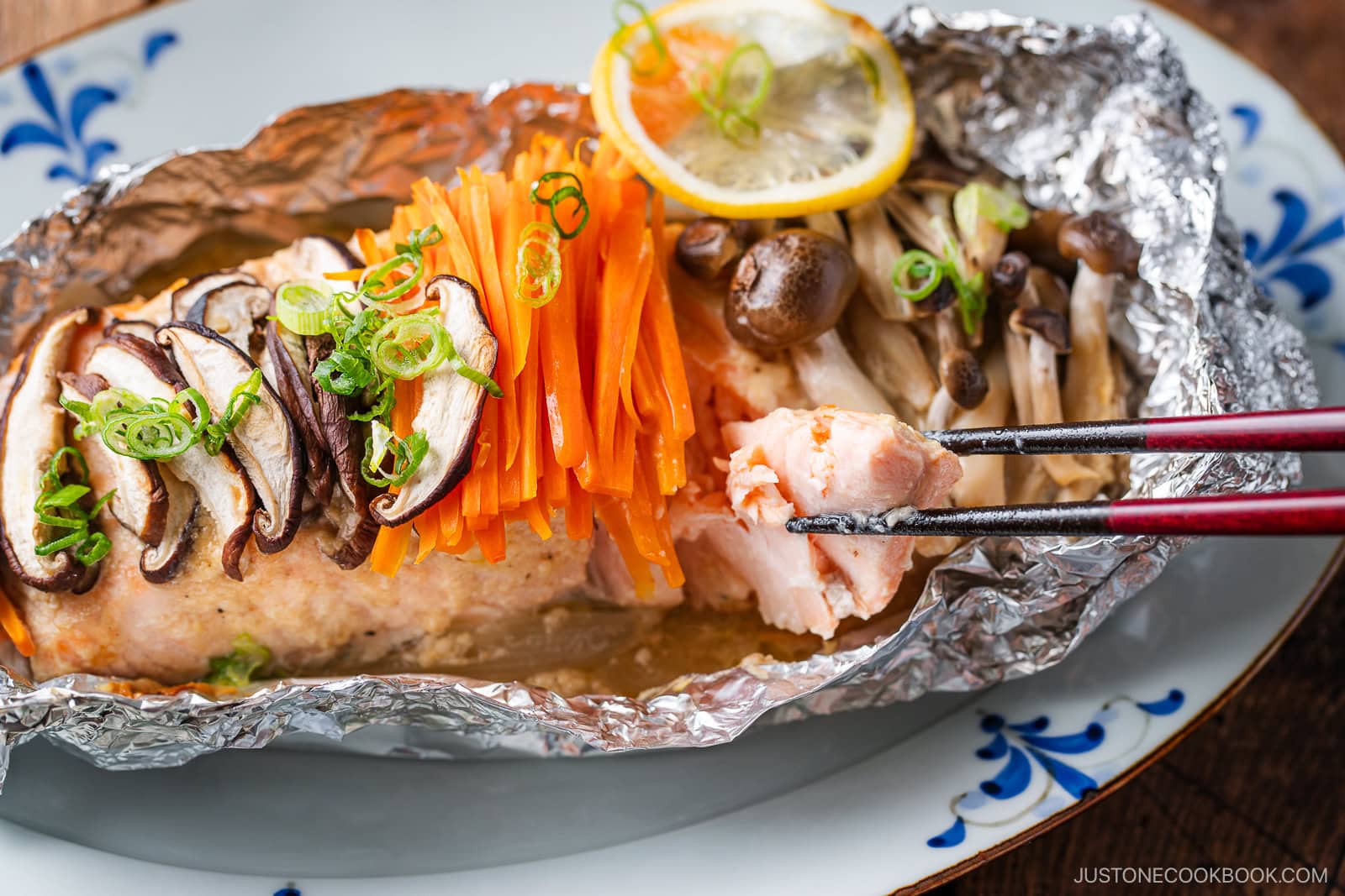 Miso salmon in foil baked to perfection, topped with sliced mushrooms, julienned carrots, green onions, and a lemon slice. A pair of chopsticks holds a piece of salmon over a decorative plate.