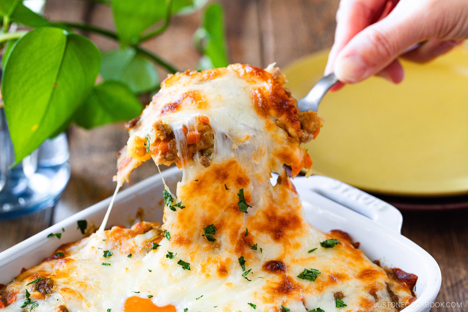 A hand lifts a cheesy, golden-brown Yaki Keema Curry casserole from a white dish with a serving spoon. Melted cheese stretches as the serving is pulled away, and chopped herbs garnish the top. A green plant and yellow plate are in the background.