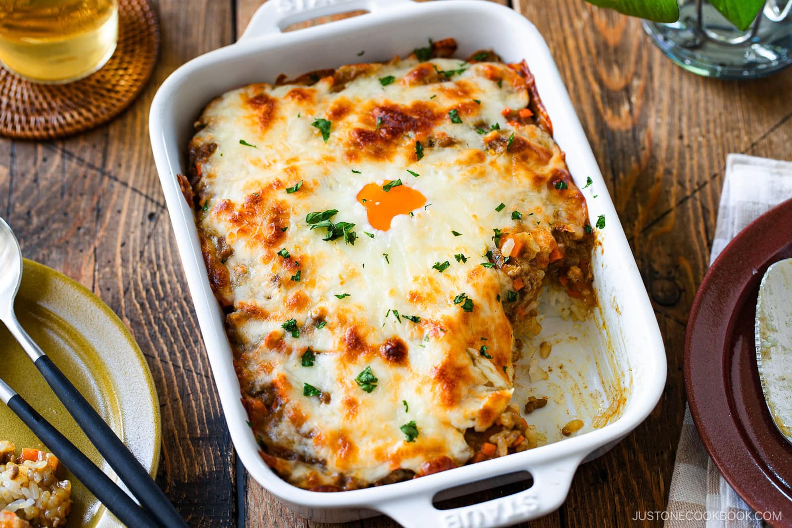 A white baking dish filled with cheesy, golden-brown Yaki Keema Curry casserole topped with fresh herbs, with one serving removed; the casserole sits on a wooden table next to a fork, plate, and drink.