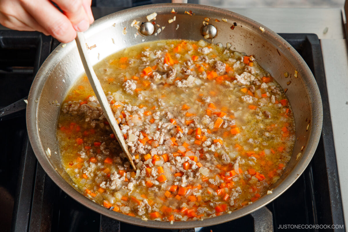 A hand stirs a savory mixture for Yaki Keema Curry—ground meat, diced carrots, and onions cooking in broth in a large stainless steel pan on a stovetop.
