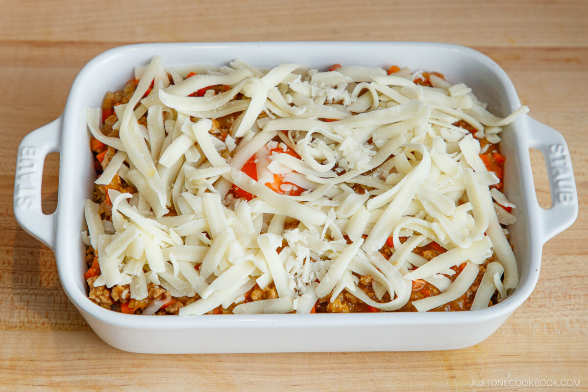 A white rectangular baking dish filled with Yaki Keema Curry casserole, topped with a generous layer of shredded cheese, placed on a wooden surface.