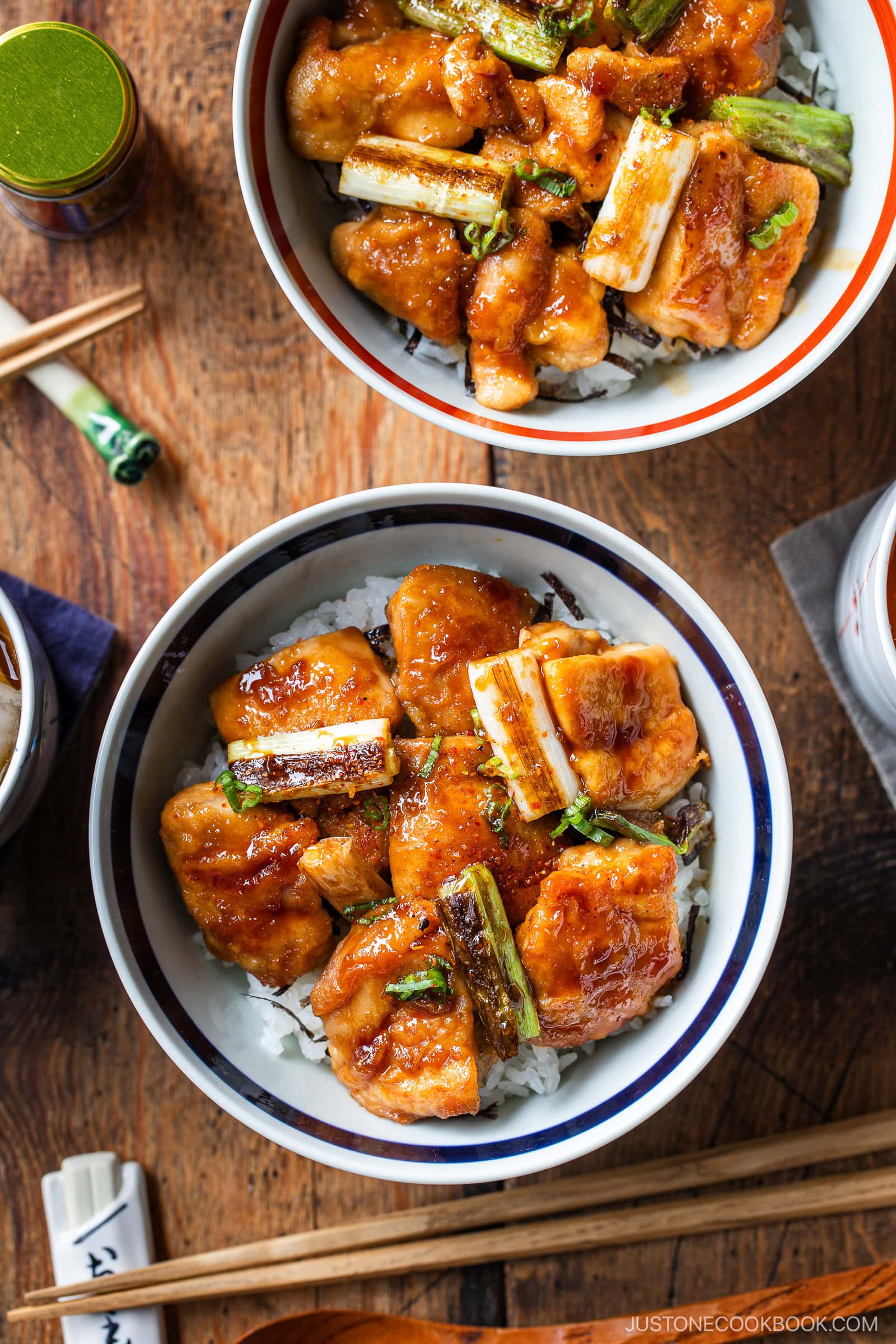 Two bowls of yakitori don—rice topped with glazed grilled chicken pieces and charred green onions—are served with chopsticks on a wooden table. The food appears golden and appetizing.