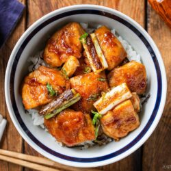 A bowl of rice topped with glazed grilled chicken pieces and scallions, this yakitori don is served with chopsticks on a wooden table. A cup and a sauce container are partially visible next to the bowl.