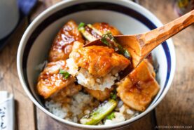 A wooden spoon lifts a piece of glazed chicken from a bowl of yakitori don, with rice, vegetables, and green peppers. The food looks saucy and freshly cooked.