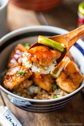 A wooden spoon holds a bite of yakitori don—glazed chicken, rice, and green onion—above a bowl filled with more saucy chicken and rice. The dish appears fresh and is garnished with chopped green onions.